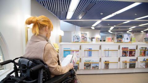 Henkilö lukee lehteä pyörätuolissa istuen kirjaston lukusalissa.