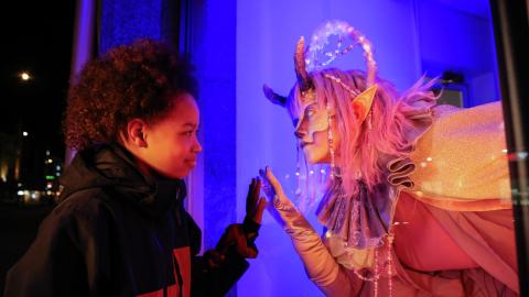 A child and a fantasy character with magical lights and horns are touching the window that separates them at the same point.