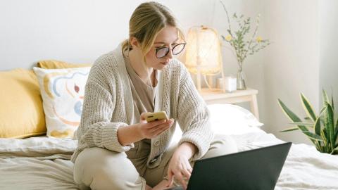 Nuori naishenkilö istuu sängyllä tietokoneen äärellä ja hänellä on kädessään puhelin.