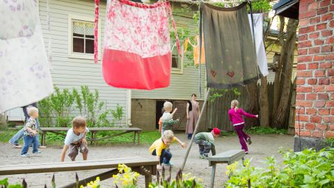 Lapset leikkivät museon pihamaalla. Etualalla narulla roikkuu pyykkiä, taustalla vaalea puutalo.