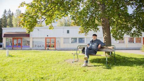 Vaalea matala talo. Sen edustalla puu ja puun alla penkillä ihminen istumassa.