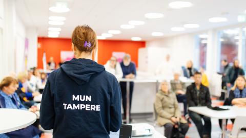 A city employee wearing a Mun Tampere hoodie is in front of the audience talking to them.