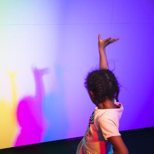 Child holding one hand up, in front of child is colorful wall.