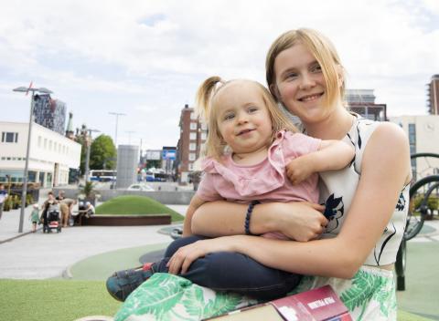 A child and a young person are sitting in a playground. A toddler is in the young person&#039;s lap.