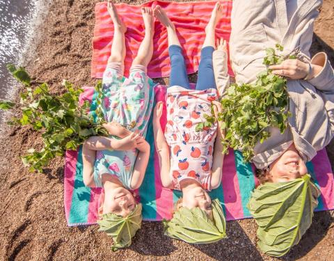 Kolme lasta makaa vierekkäin rantahietikolla suuret raparperinlehdet kasvoillaan ja saunavihdat kädessä. 