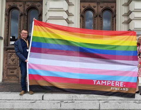 Two people are standing on the steps of Tampere Town Hall. They are displaying a large Tampere pride flag with 13 colour stripes.