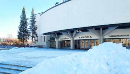 Sampola-rakennus. Vieressä on havupuita, ja etualalla on lumikinoksia.