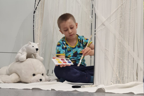 Lapsi istuu lattialla valkoisessa majassa ja maalaa majan valkoisista langoista tehtyä seinää eri värisillä vesiväreillä.