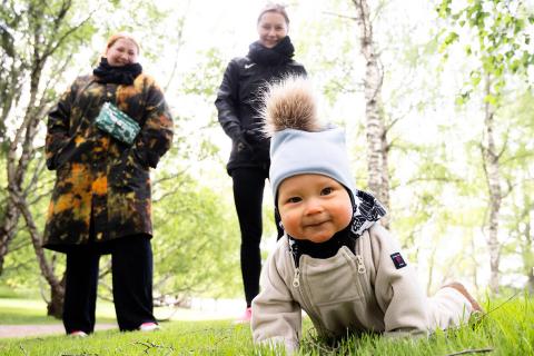 Pieni lapsi ryömii kuvan etualalla nurmella päällään haalarit ja tupsupipo ja kaksi aikuista seisoo lapset takana.